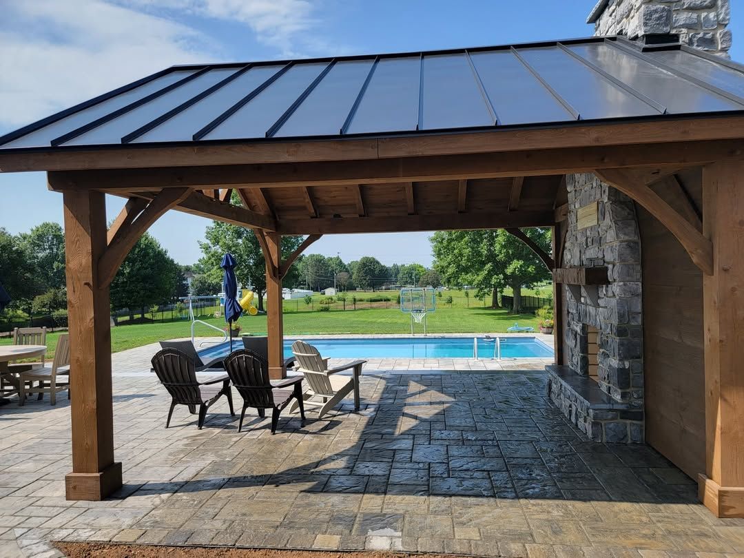 Jesse James Hardscaping - Outdoor pavilion with fireplace, overlooking a pool and yard; wooden structure, chairs.