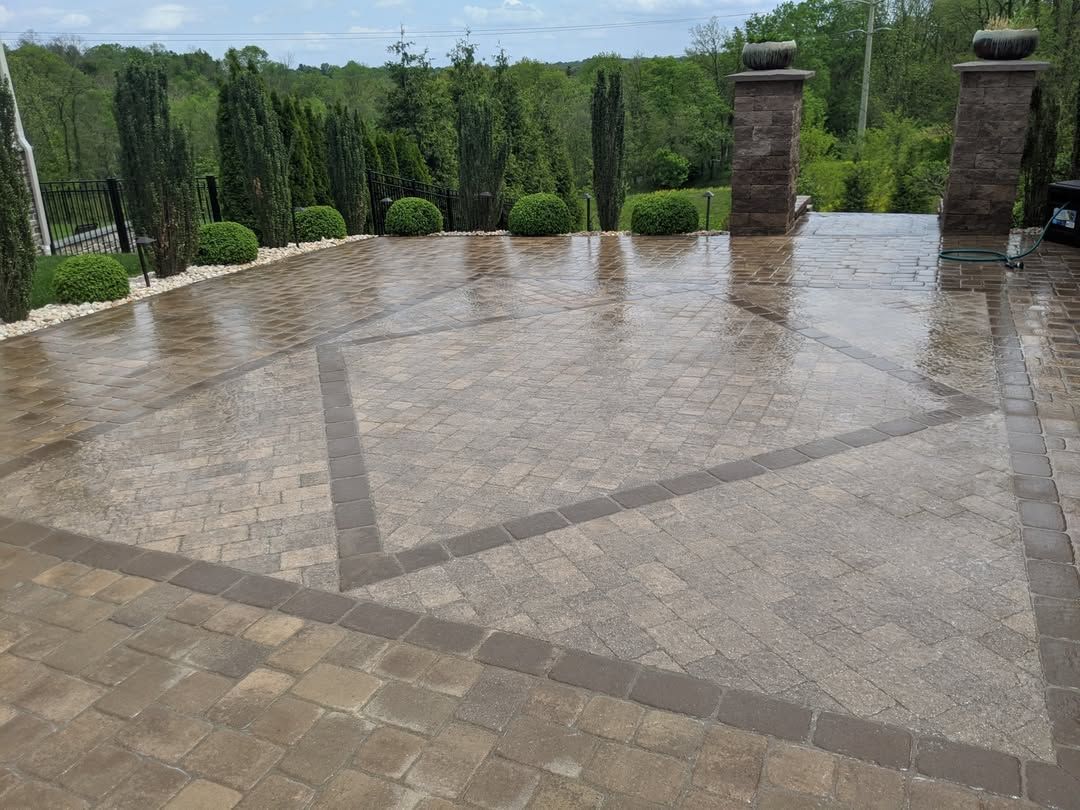 Jesse James Hardscaping - Patio with wet pavers and stone columns, geometric design, and lush green background.