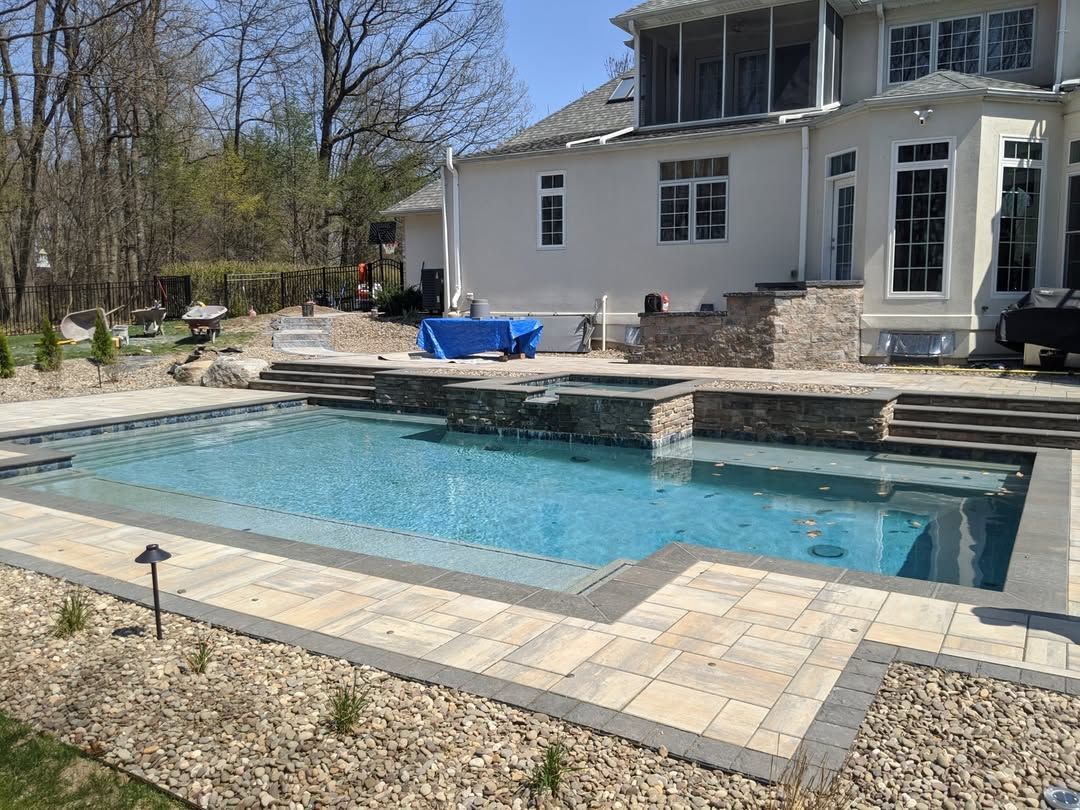 Jesse James Hardscaping - A backyard pool with stone and brick surround, next to a large house. Sunny day.