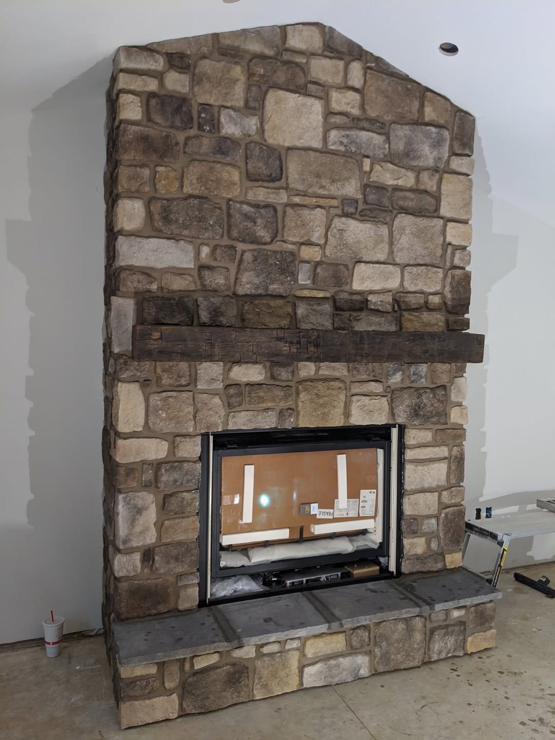 Jesse James Hardscaping - Stone fireplace with a dark wood mantel, installed in a room with unfinished walls.