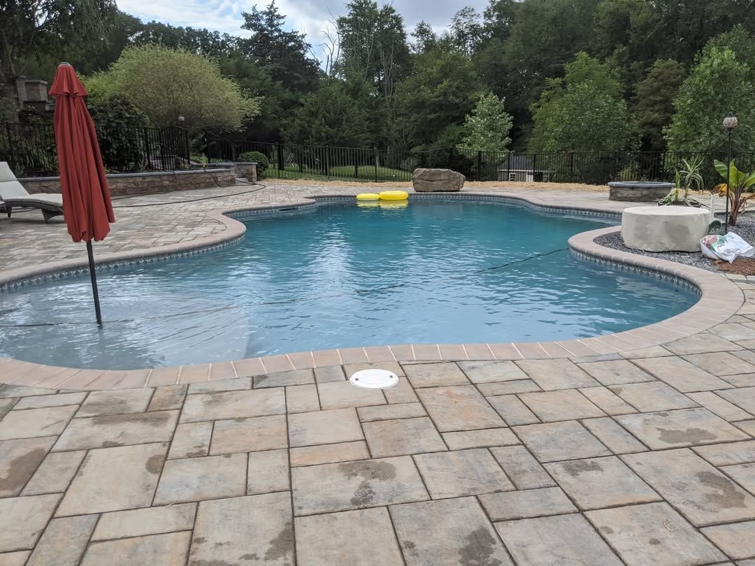 Jesse James Hardscaping - Backyard pool with paved patio, red umbrella, and trees in the background. Two yellow floats in the water.