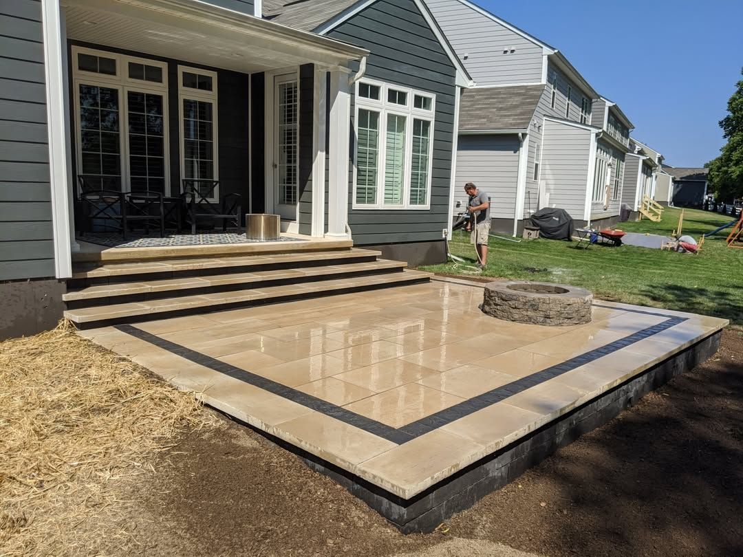Jesse James Hardscaping - Stone patio with steps, fire pit, house exterior. A person is working on the lawn. Sunny day.