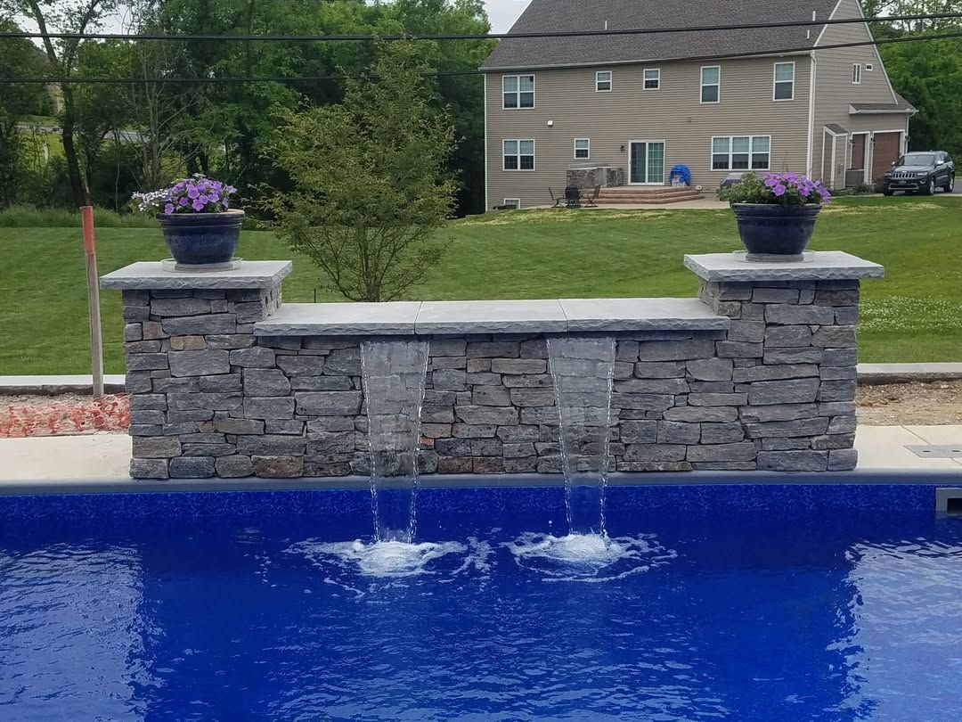 Jesse James Hardscaping - A stone waterfall feature with two water spouts flows into a blue-tiled pool, topped by flower pots.