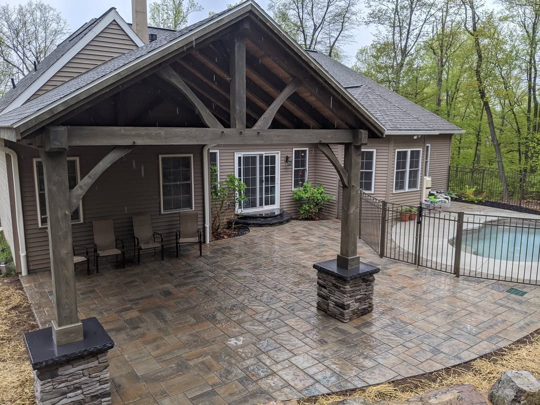 Jesse James Hardscaping - Patio with timber frame pergola, brick pavers, house, pool.  Brown tones. Cloudy day.