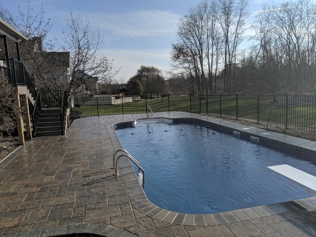 Pool with stone patio, diving board, and metal railing. A house with stairs is on the left. Sunny day.