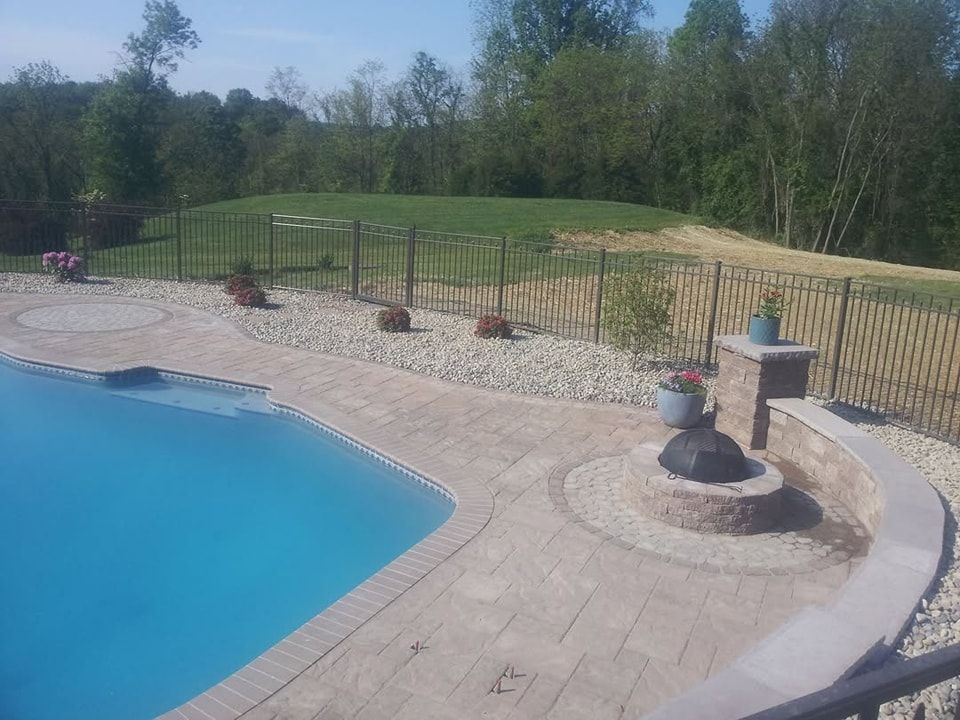 Jesse James Landscaping - Swimming pool with a fire pit area, surrounded by rocks, a fence, and a green grassy backdrop.