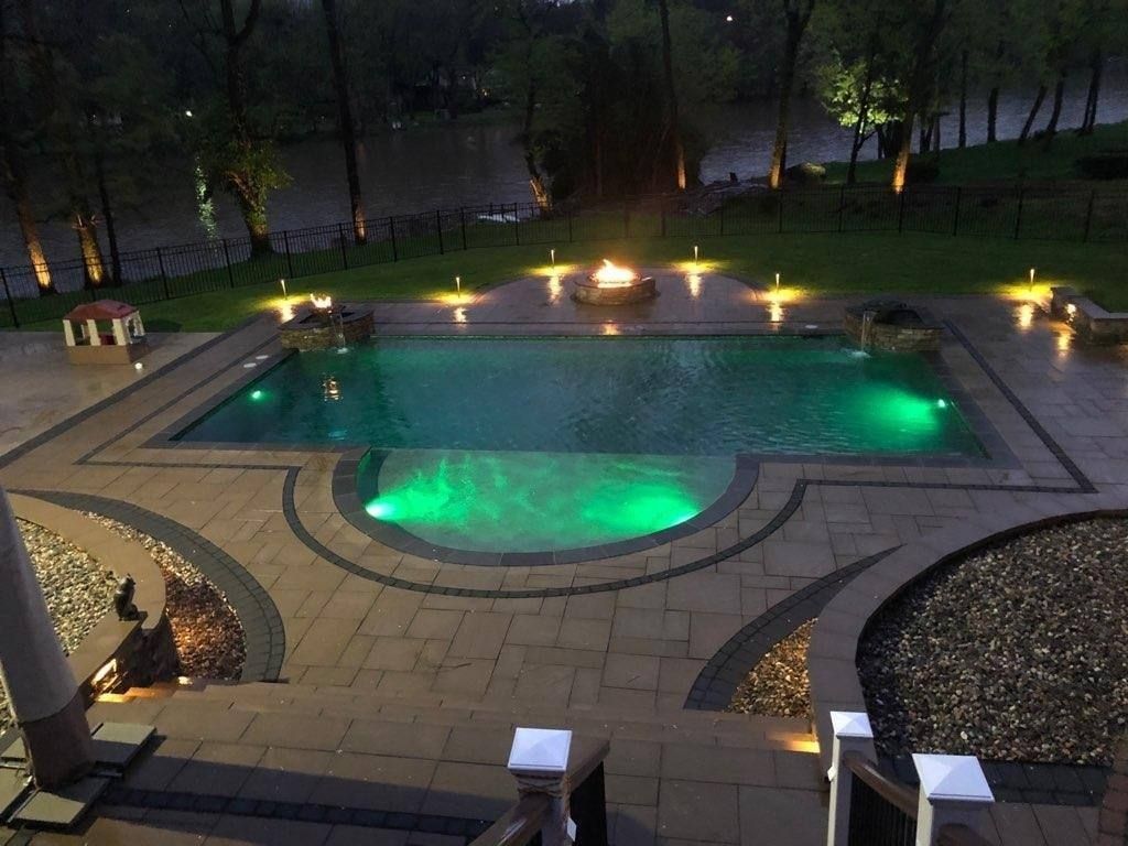 Jesse James Landscaping - Nighttime view of a pool with green lights, fire pit, and surrounding patio with river in the background.