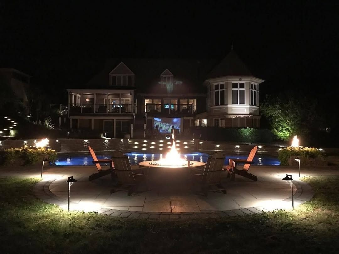 Jesse James Landscaping - Nighttime view of a large house with a fire pit and pool, surrounded by lights.