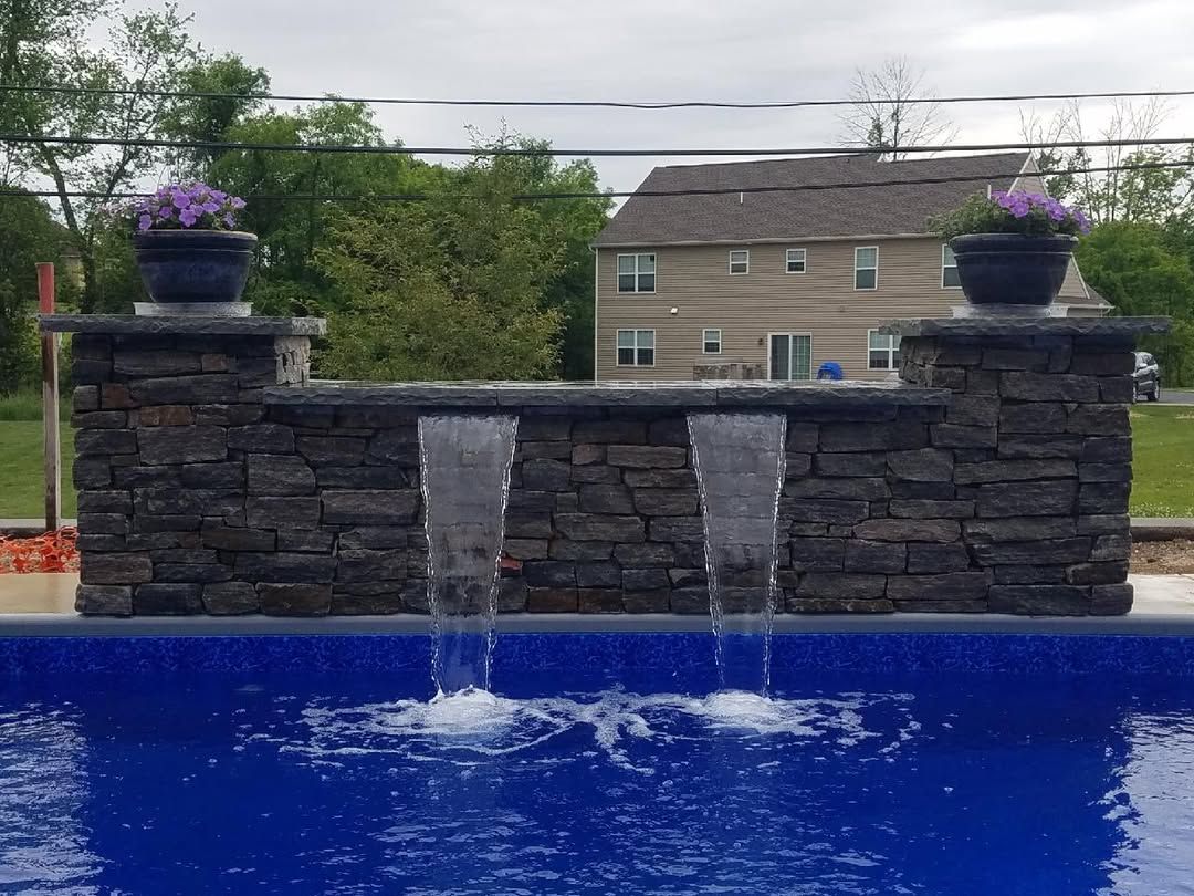 Jesse James Landscaping - Water feature with dark stone wall and two waterfalls spilling into a blue-tiled swimming pool. Purple flowers in pots on top.