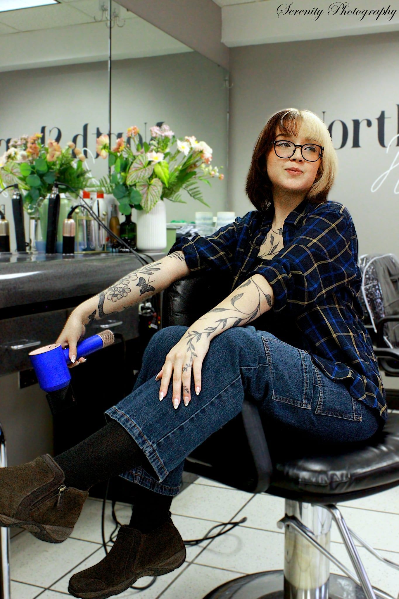 Person seated in a salon chair, holding a blue drink, with tattooed arms and a tiled floor in view.