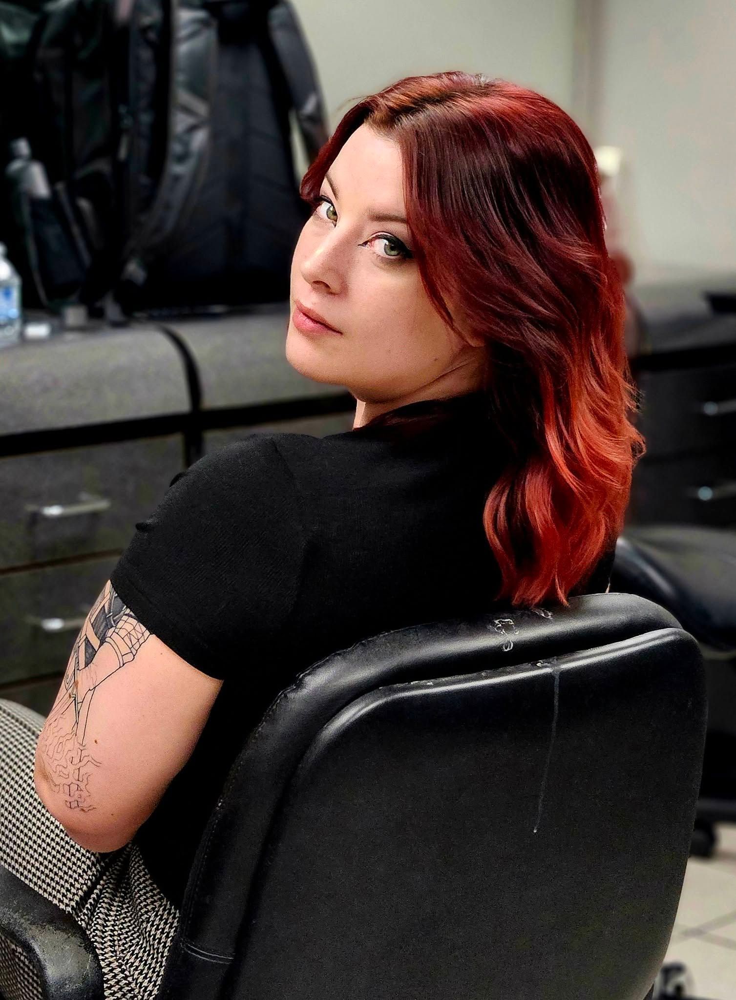 Person with red hair sitting in a black chair, looking back over their shoulder in a dim indoor setting