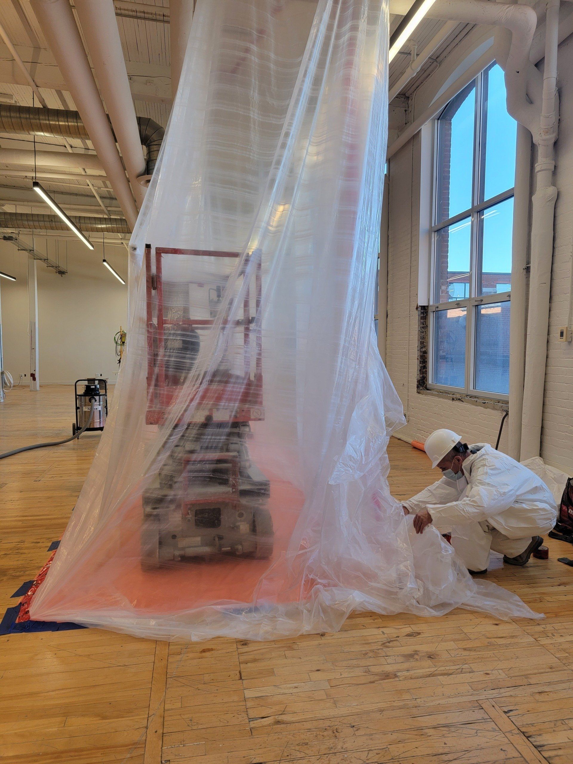Un ouvrier en combinaison de protection scelle un confinement en plastique autour d'une machine, près d'une fenêtre dans un bâtiment.