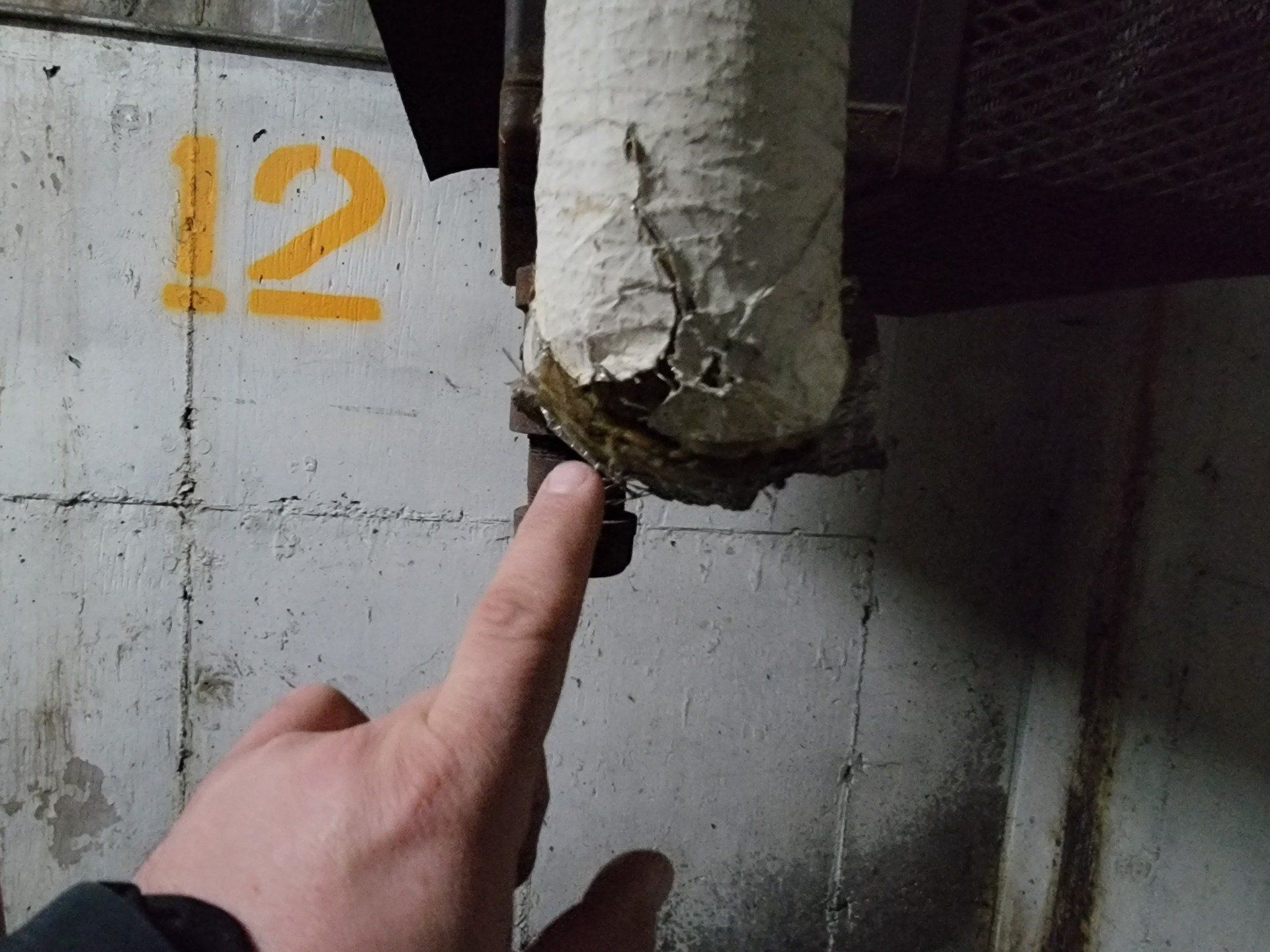 Main pointant vers un tuyau endommagé et isolé fixé à un mur en béton marqué « 12 ».