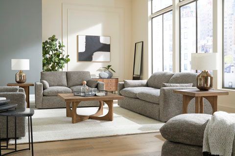 A modern living room with gray sofas, wooden tables, and an abstract painting. Natural light fills the space.