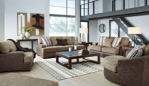 Living room with brown sofas, a patterned rug, and abstract artwork, against a backdrop of large windows and a staircase.