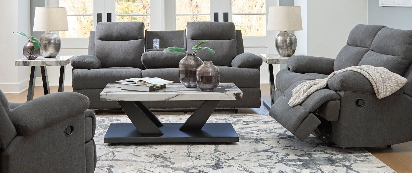 A living room with gray reclining sofas and chairs, a marble-topped coffee table, and side tables with lamps.