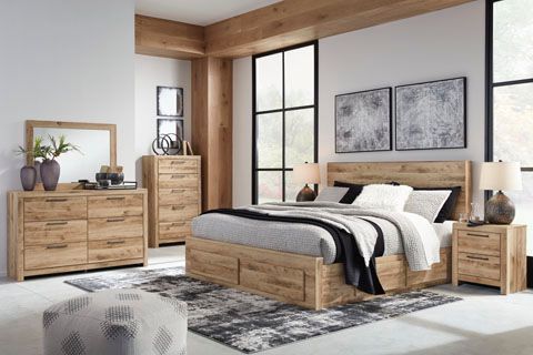 A bedroom with light-wood furniture: bed, dresser, chest, nightstand, and mirror. A textured rug sits on the floor.