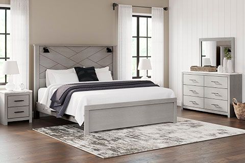 A bedroom with gray furniture: bed, nightstand, dresser, and mirror. White bedding, rug, and curtains.