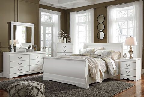 White bedroom set with sleigh bed, dresser, nightstand, chest, and mirror, on a gray rug, with white curtains and dark walls.