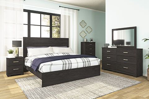 Bedroom set with a dark wood finish, including a bed, nightstand, dresser, chest, and mirror. The room has a light blue wall and white curtains.