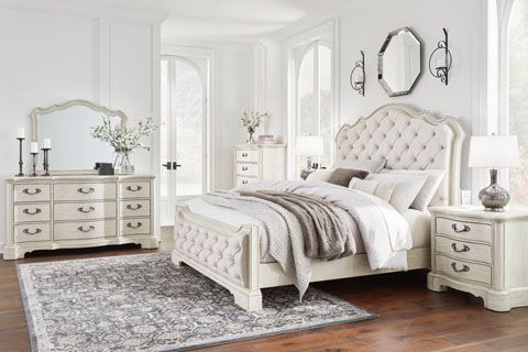 A bedroom set with a tufted bed, dresser, nightstand, and mirror. The room is light-colored and features decorative accents.