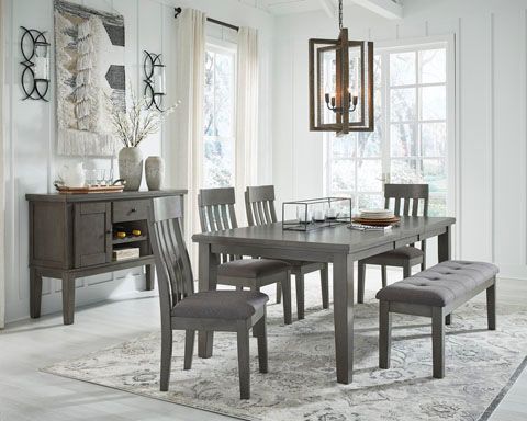 A gray dining room set includes a table, chairs, and a bench, with a matching buffet against a white wall, lit by a chandelier.