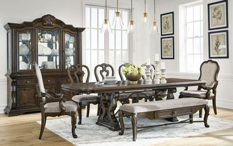 Formal dining room set with a dark wooden table, ornate chairs, a buffet, and a hutch, all in a bright room with light fixtures and framed artwork.