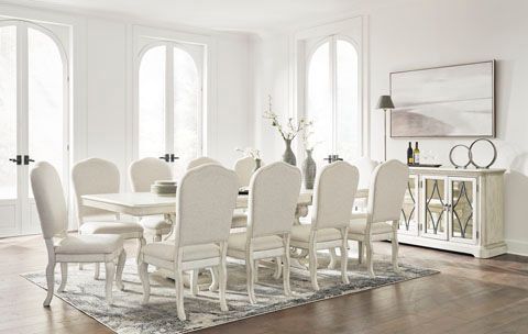 A bright dining room with a white table and ten white chairs, a buffet, and large windows.