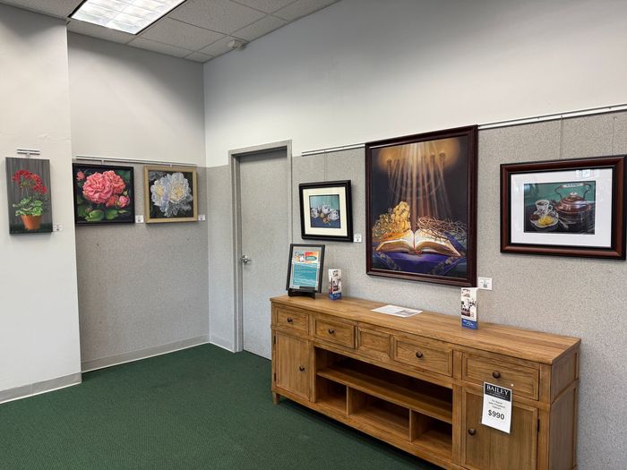 Art gallery space featuring framed paintings on display above a wooden cabinet and along the wall. Green carpeted floor.