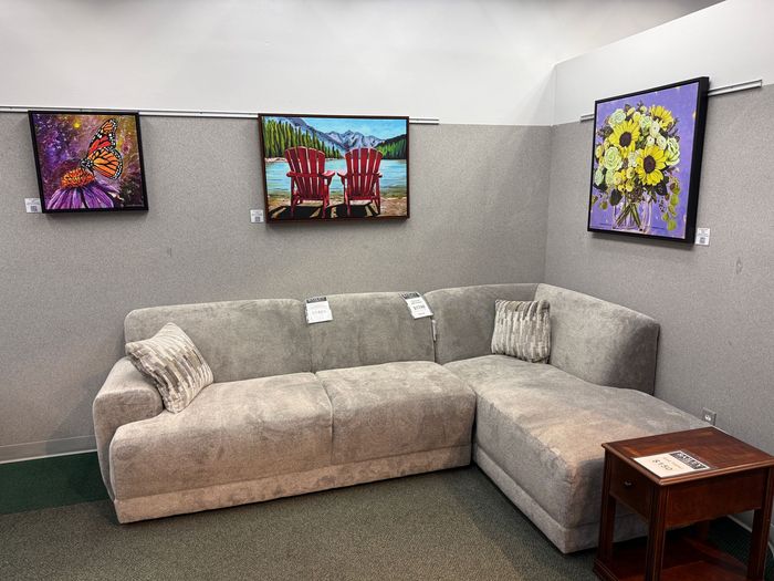 A sectional sofa in a showroom setting with artwork on the wall. The artwork includes a butterfly, lake scene, and sunflowers.