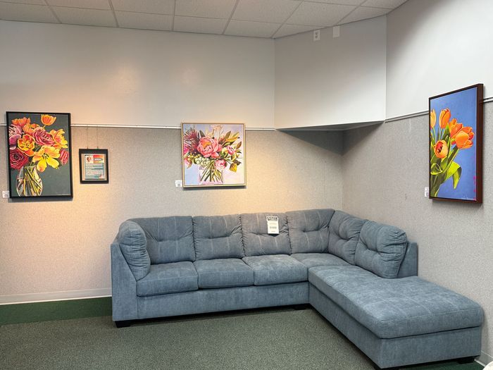 A small gallery setting with a blue sectional sofa, showcasing floral paintings on the walls.
