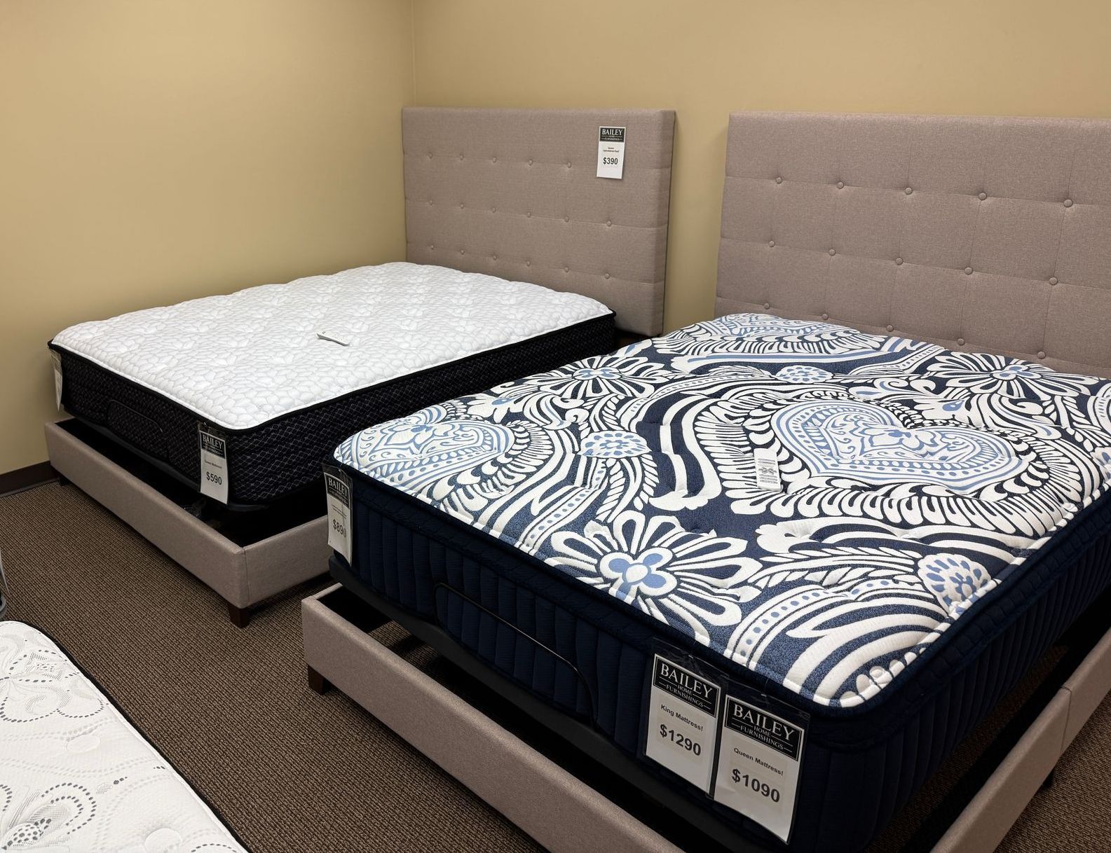 Two mattresses with upholstered headboards in a showroom. One has a white top, the other a dark blue and white pattern.