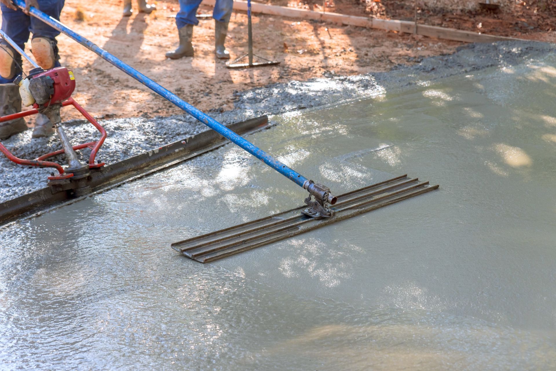 Workers smoothing wet concrete with a long blue-handled screed on a construction site