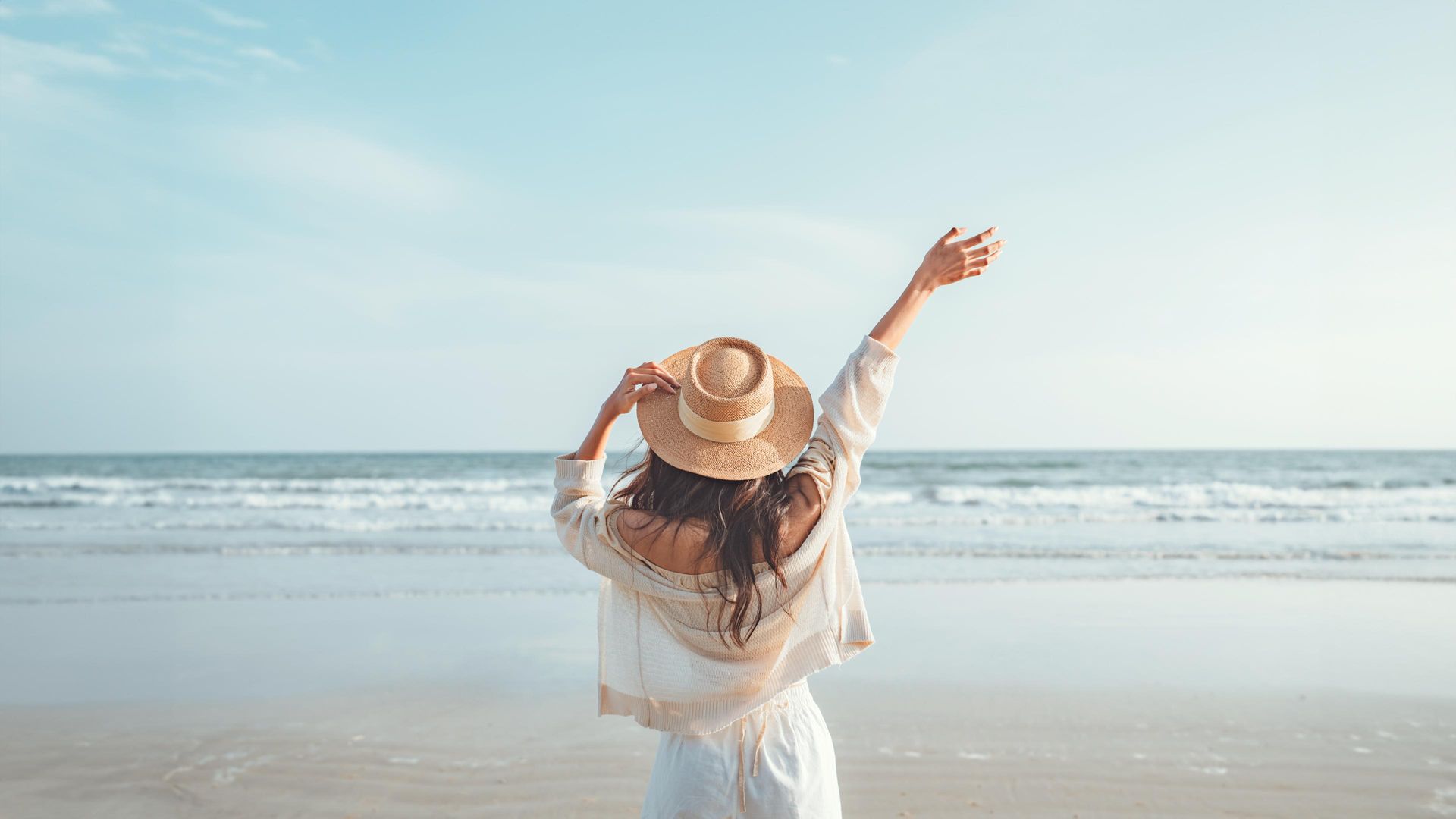 A person wearing a straw hat and white outfit stands on a sandy beach.