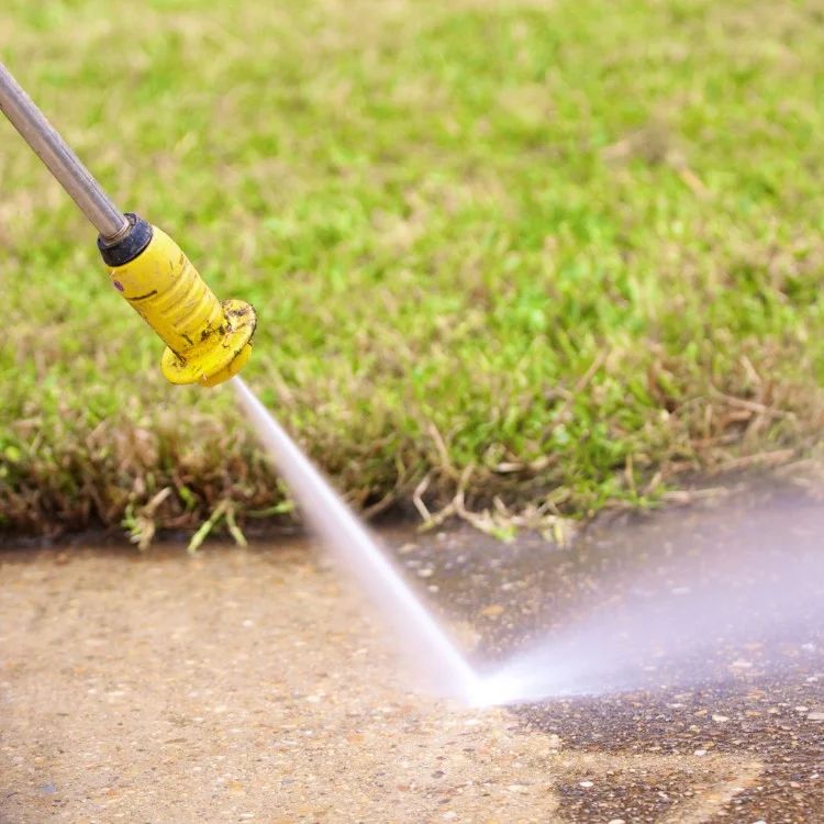 A pressure washer nozzle sprays water onto a concrete surface beside grass.
