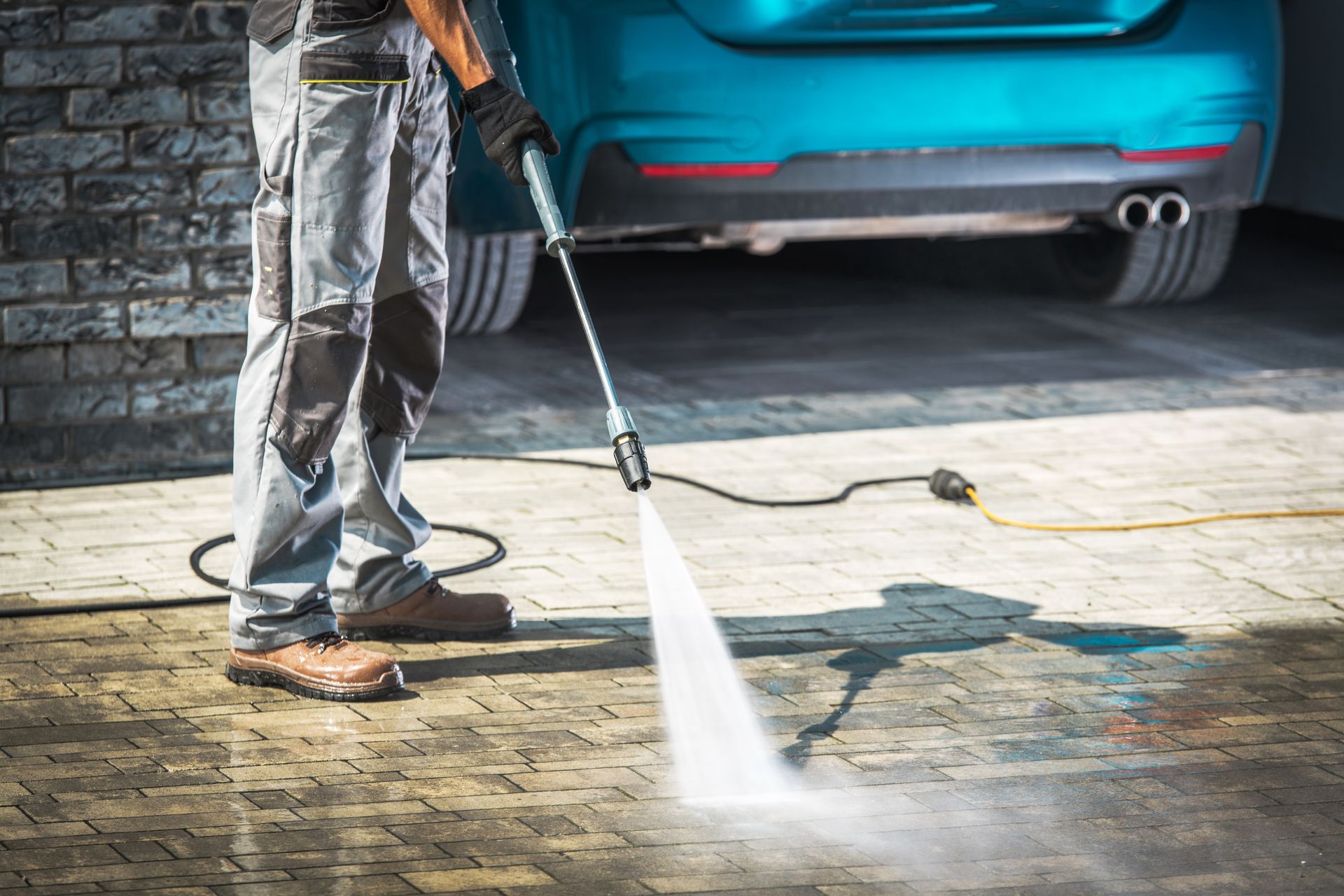 Person pressure-washing a driveway beside a blue car in a garage.