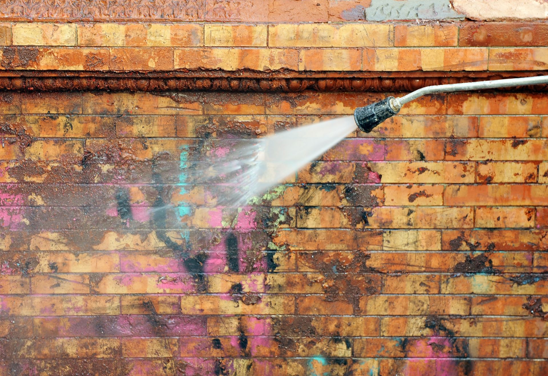 A hose sprays water on a graffiti-covered brick wall, cleaning a section with pink and blue paint.