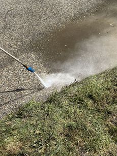 Person spraying a driveway with a blue hose nozzle, cleaning wet concrete beside grass.