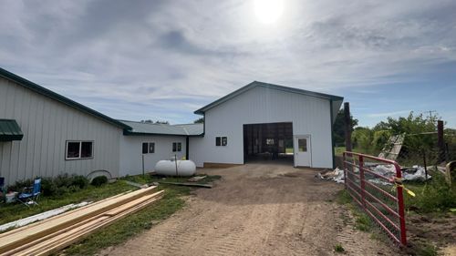 White barn with open door, green roof, gas tank, and lumber on a gravel driveway under a cloudy sky.