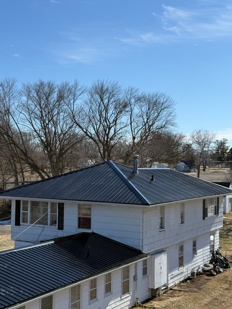 A two-story white house with a dark metal roof, set against a bright blue sky with bare trees in the background.