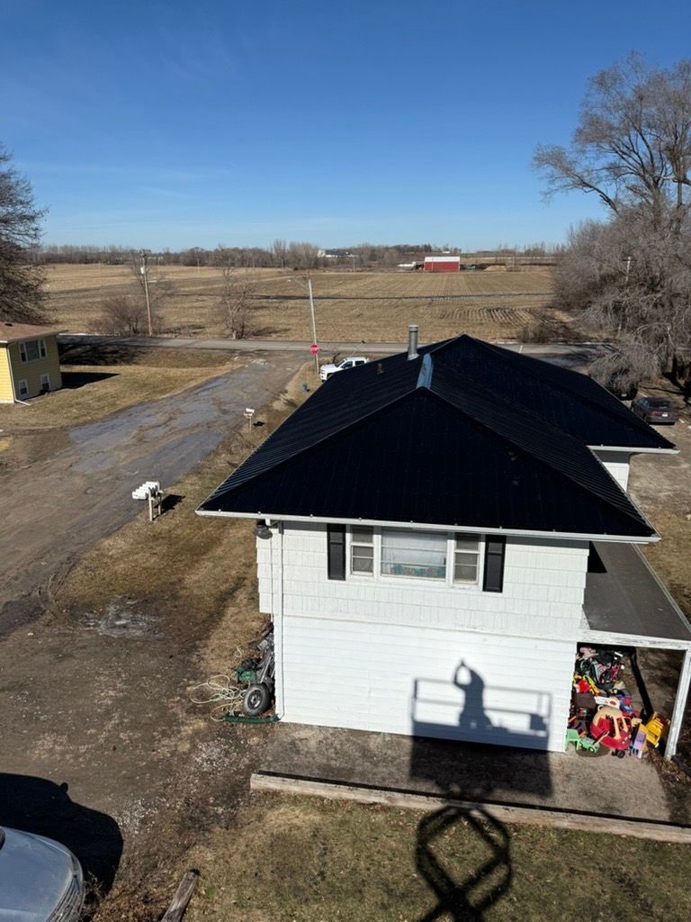 An aerial view of a white house with a new black roof, surrounded by a rural field under a clear blue sky.