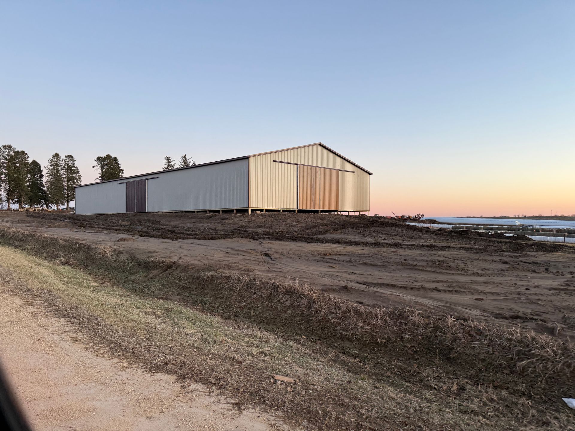 A long, light-colored metal barn stands on a cleared dirt lot at sunset under a clear sky.