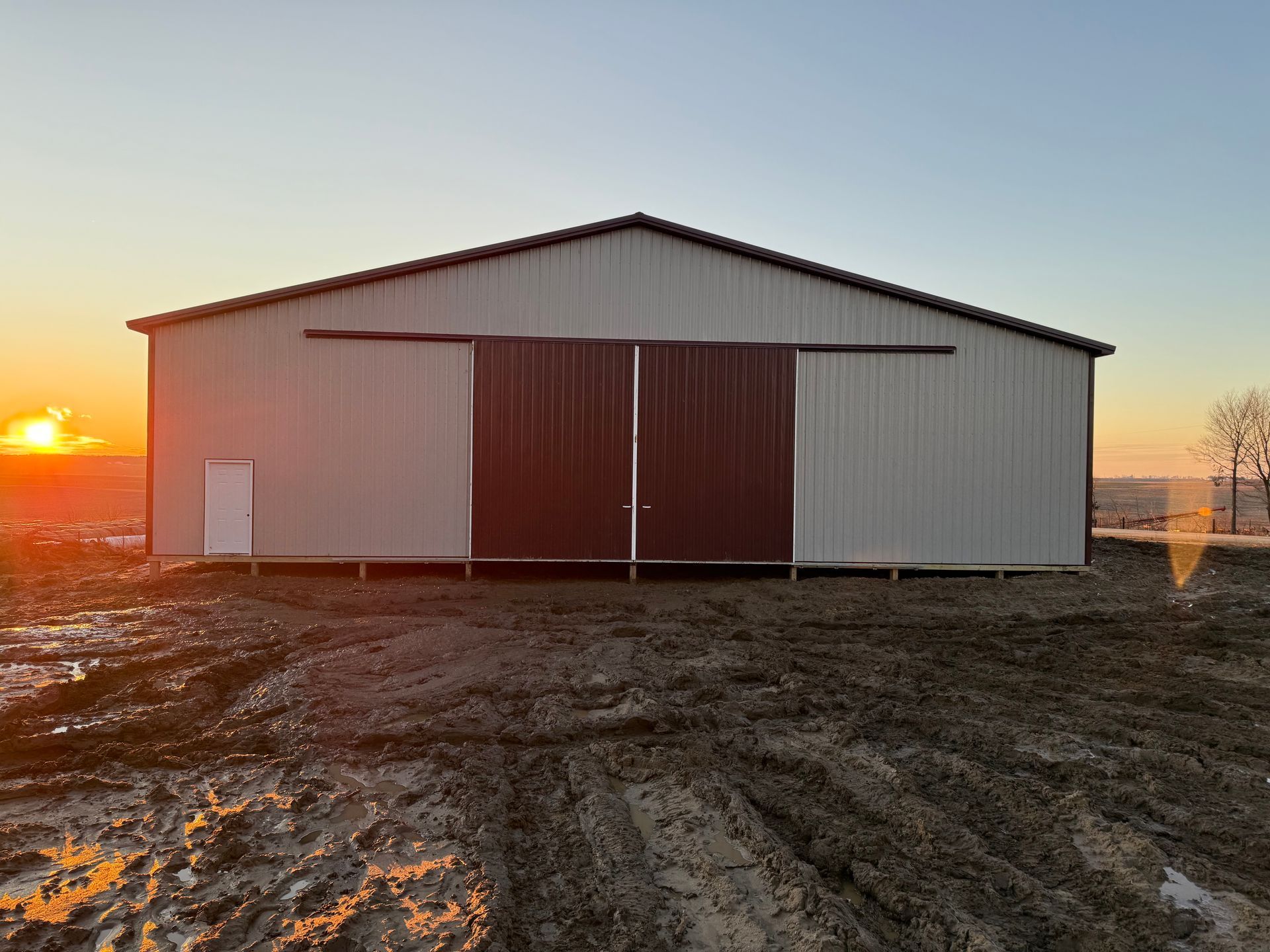 A wide, metal barn with sliding red doors and a small white side door set in a muddy, rural field at sunset.