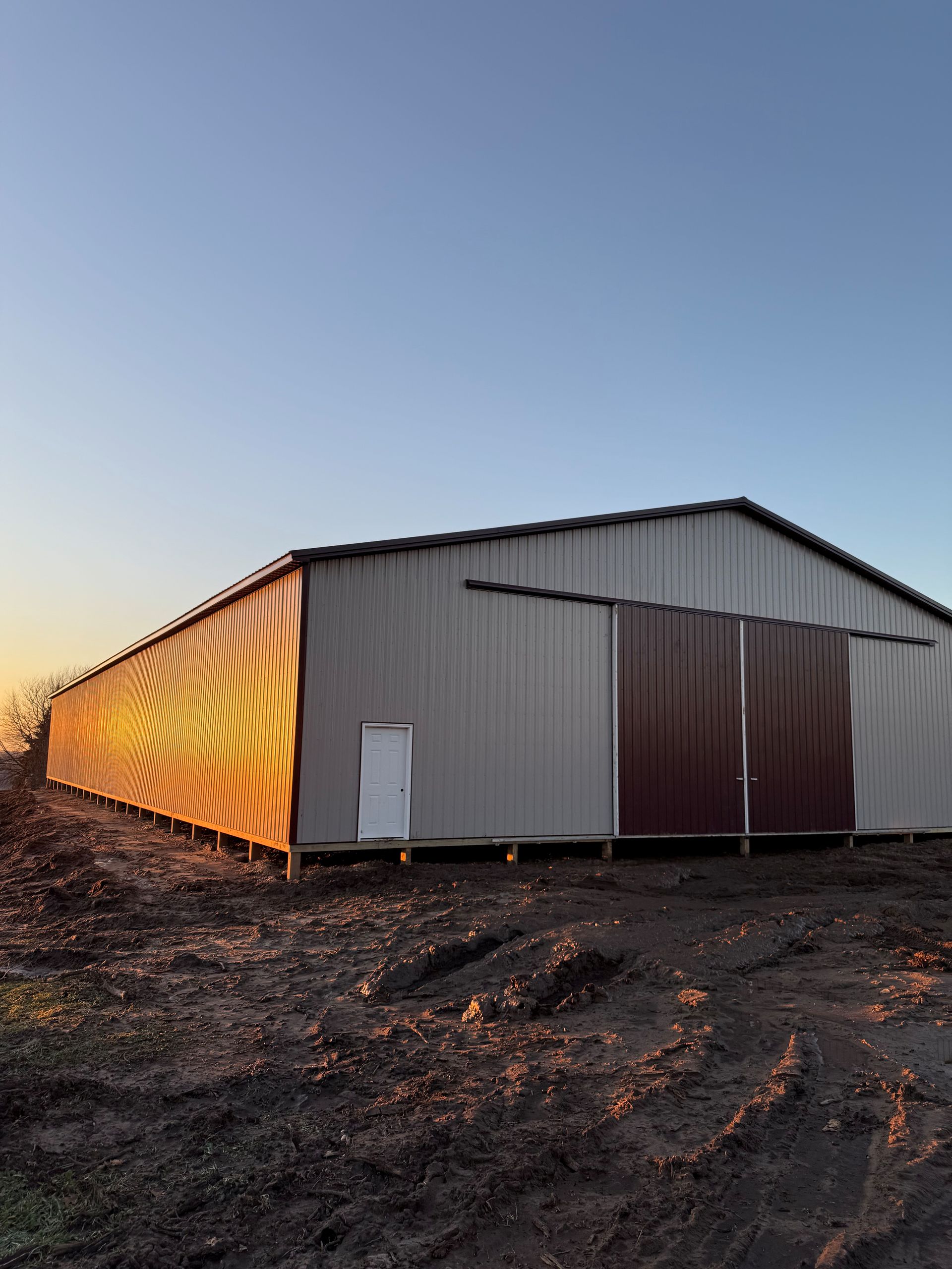 A metal agricultural building with a white door and dark sliding doors, backlit by a sunset in a muddy field.