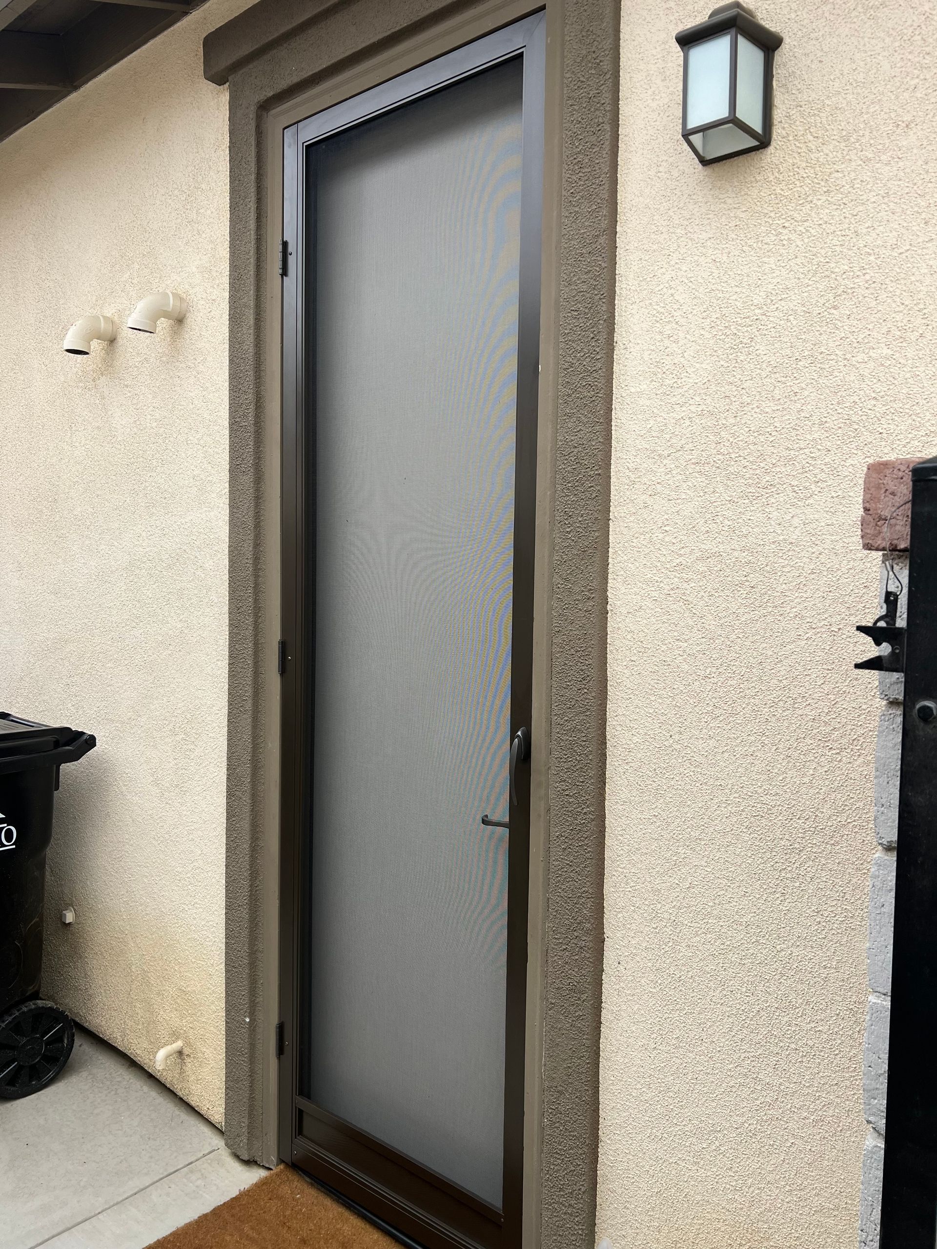 A brown screen door is set in a stucco wall. A black trash can is to the left, and a lantern is on the wall above.