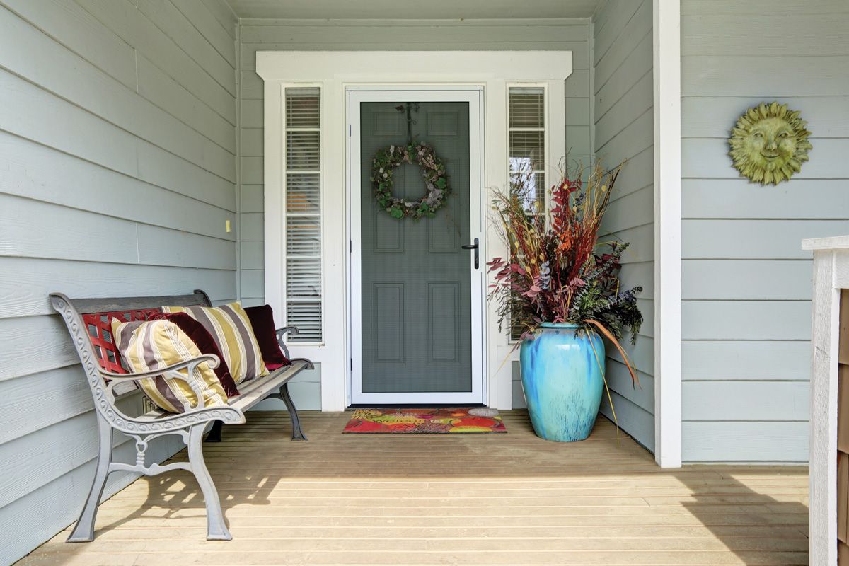 Gray front door with wreath, bench with pillows, and turquoise vase on a porch.