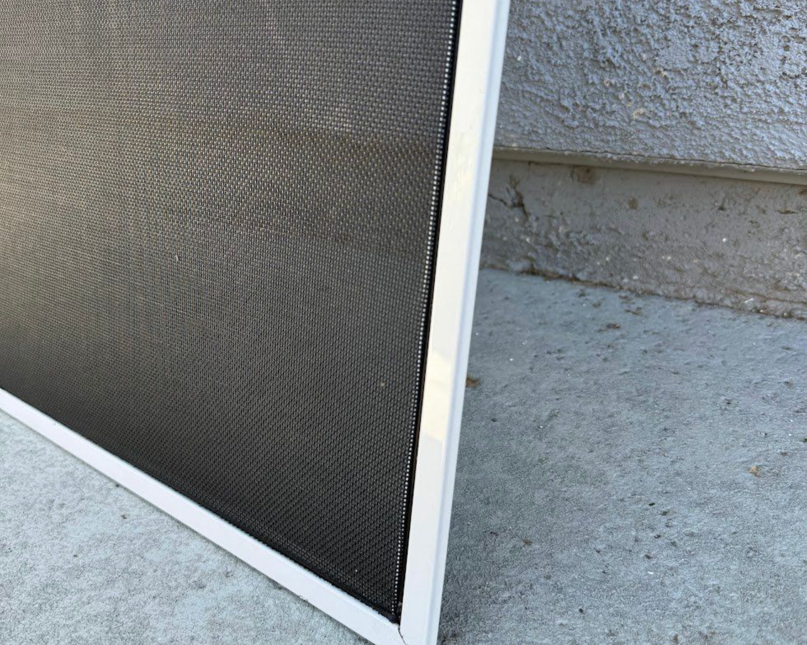 White-framed window screen with dark mesh, resting against a textured gray wall and concrete surface.