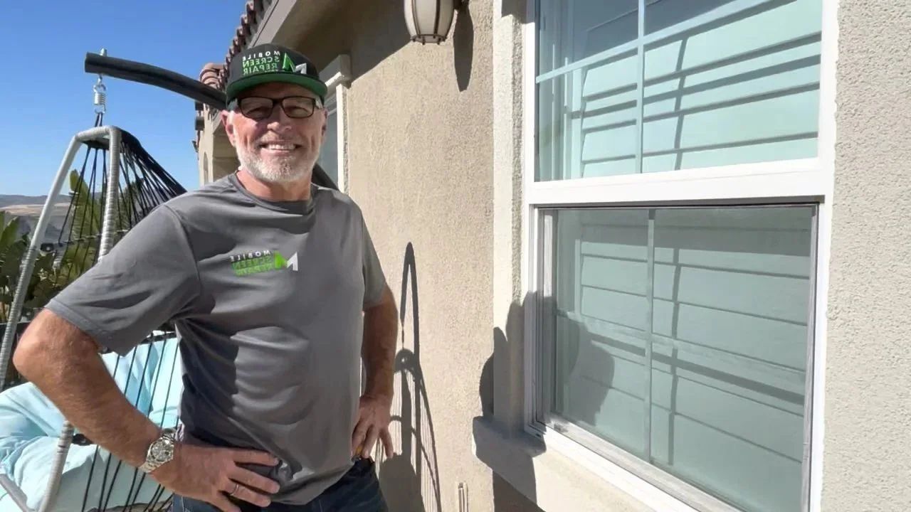 Owner of a mobile screen repair service standing beside a window with installed screen mesh, outdoors at a home.