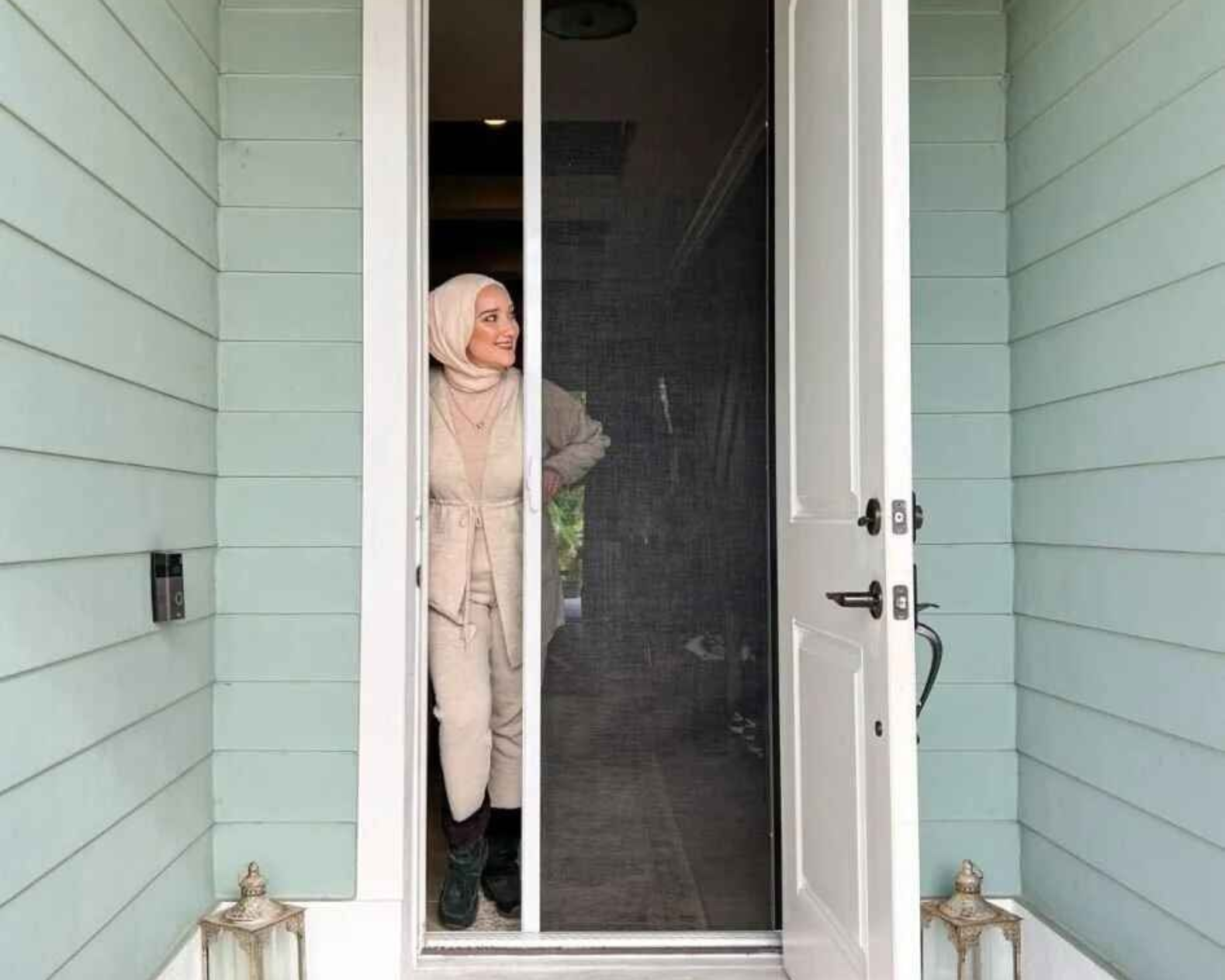 Woman in hijab stands in doorway with retractable screen door, white door, and blue siding.