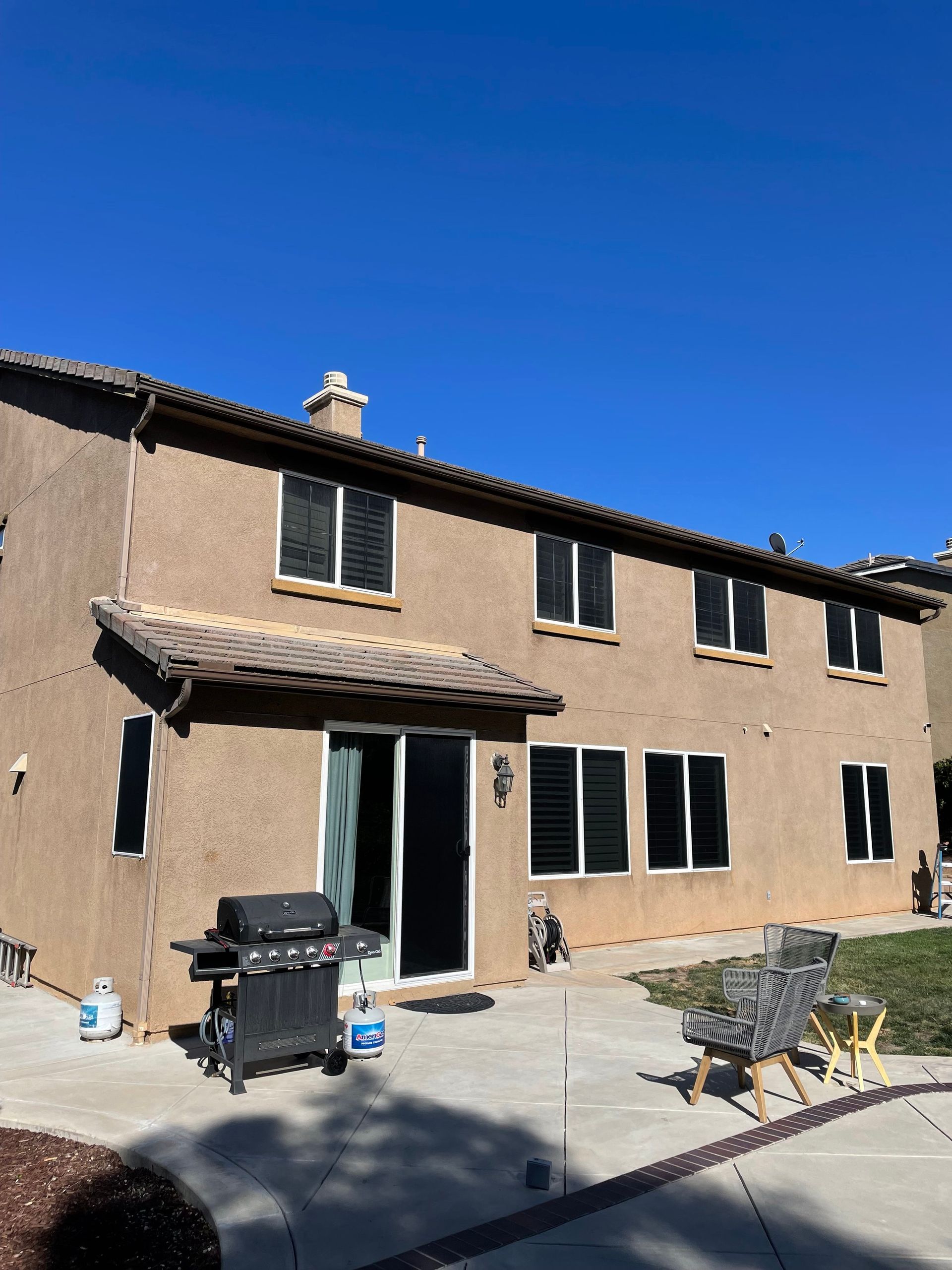 Back exterior of a two-story beige house with dark windows, sliding glass door, patio, and blue sky.